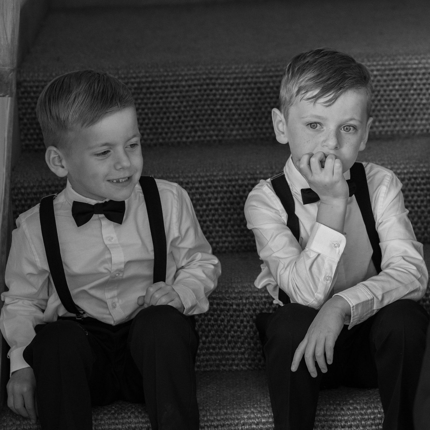 Two young boys in formal attire with bow ties and suspenders sitting together on steps in black and white.