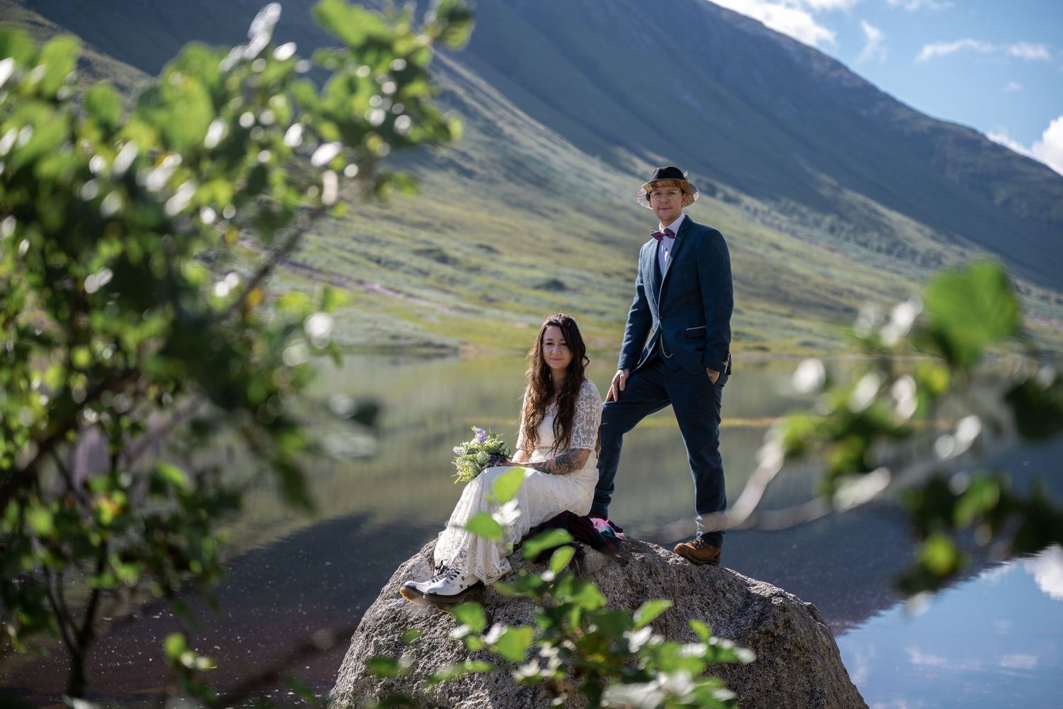 A couple poses dramatically on a rocky outcrop overlooking scenic mountain landscape.