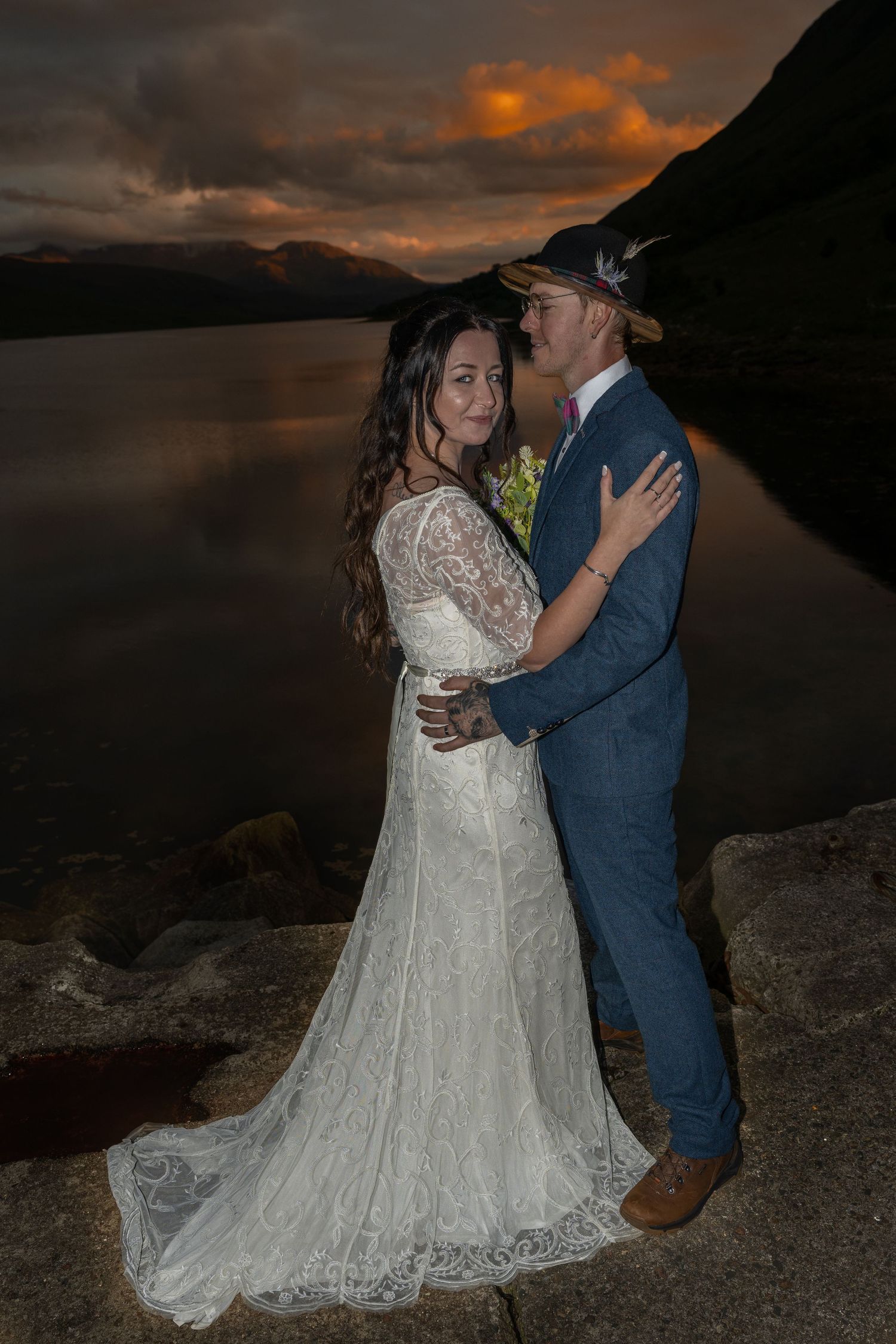 Wedding couple embraces against dramatic mountain lake sunset backdrop.