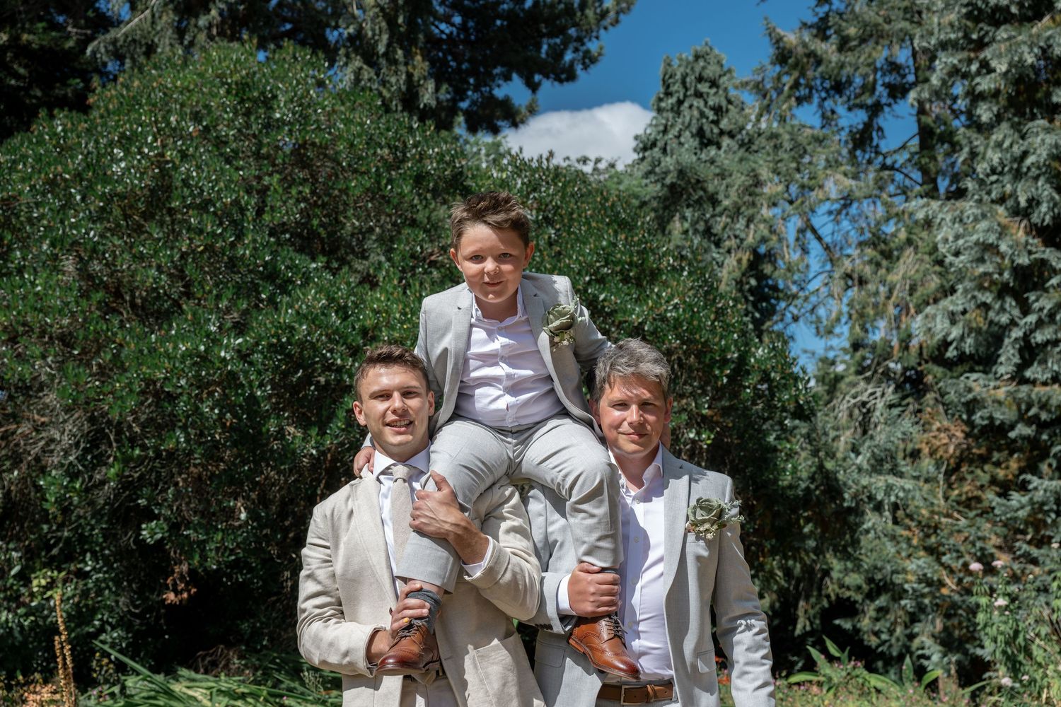 Three people in light colored suits pose playfully in a garden setting.