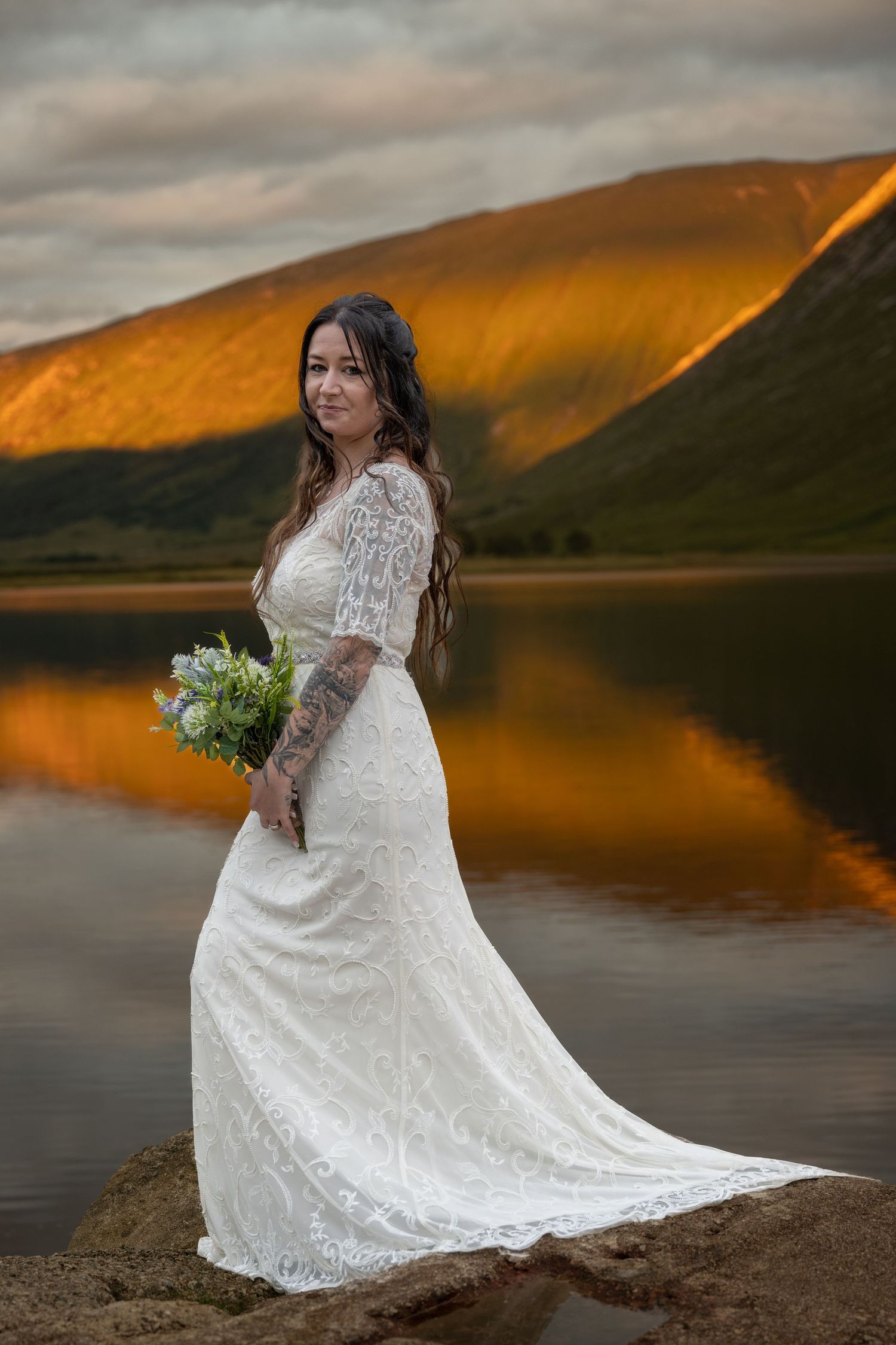 Bride in lace dress poses by mountain lake at sunset with golden light reflecting on water.