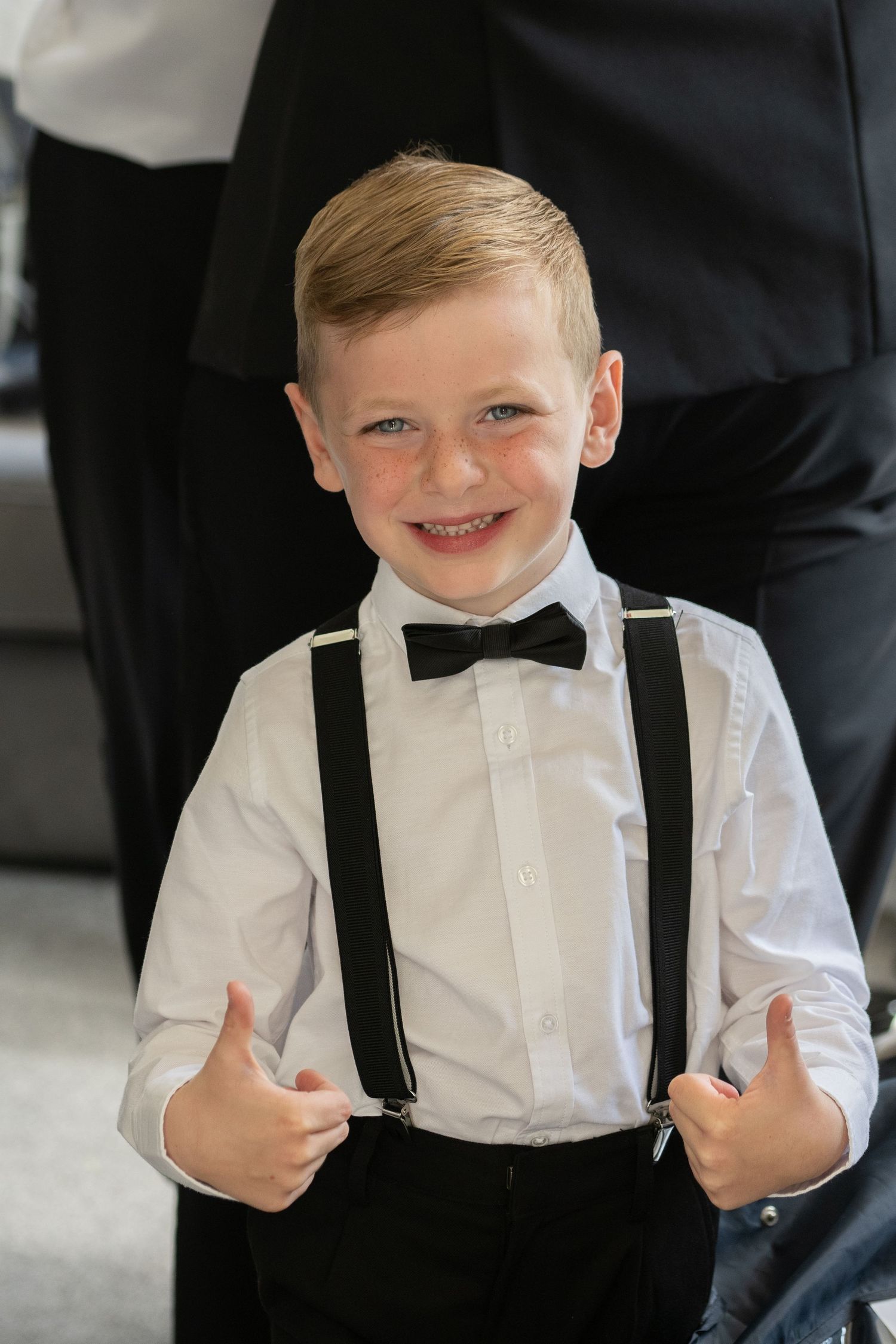 Cheerful young boy in formal attire giving thumbs up while wearing suspenders and bow tie.
