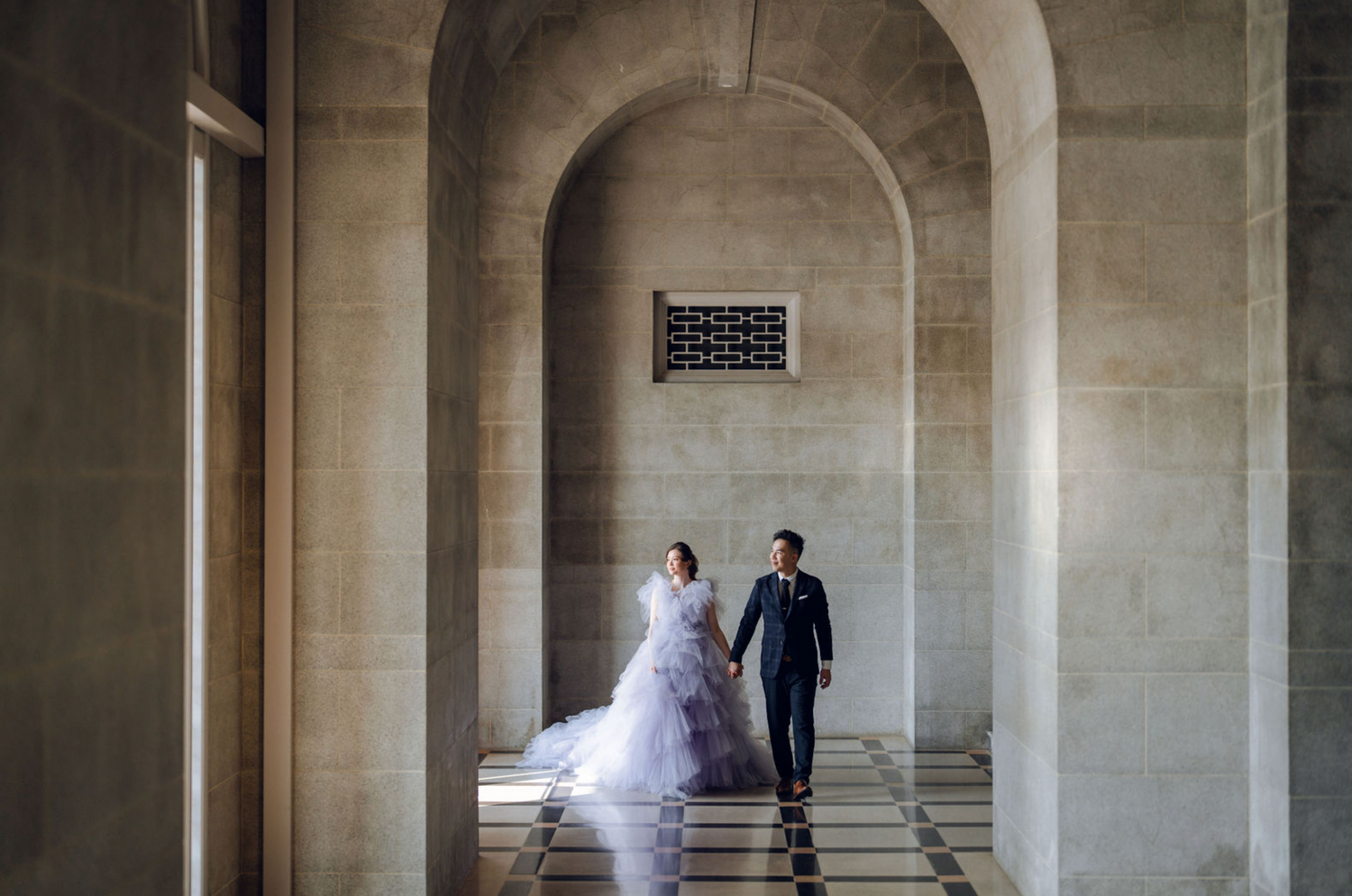 Wedding couple walks through stone archways in a dramatic architectural setting.