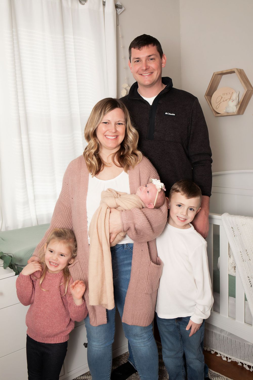 Family portrait in a cozy nursery with white curtains and geometric decor.