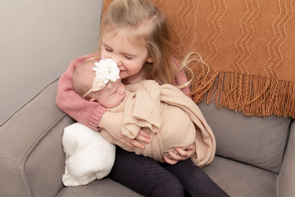 Young child cuddling a swaddled baby on a gray couch with an orange throw blanket.