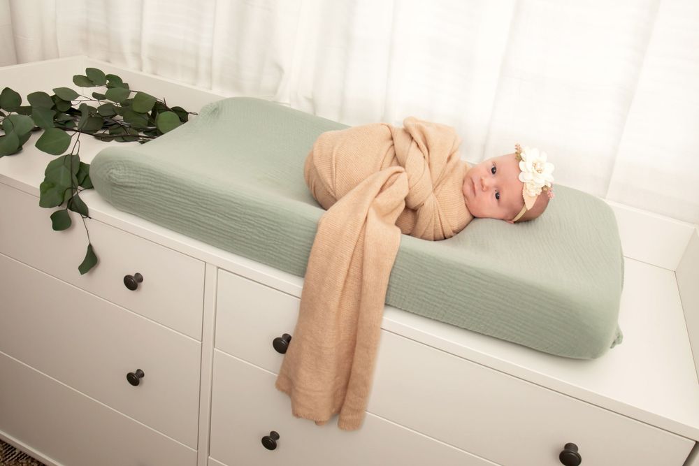 Baby wrapped in beige swaddle resting on mint green changing pad with greenery decor.
