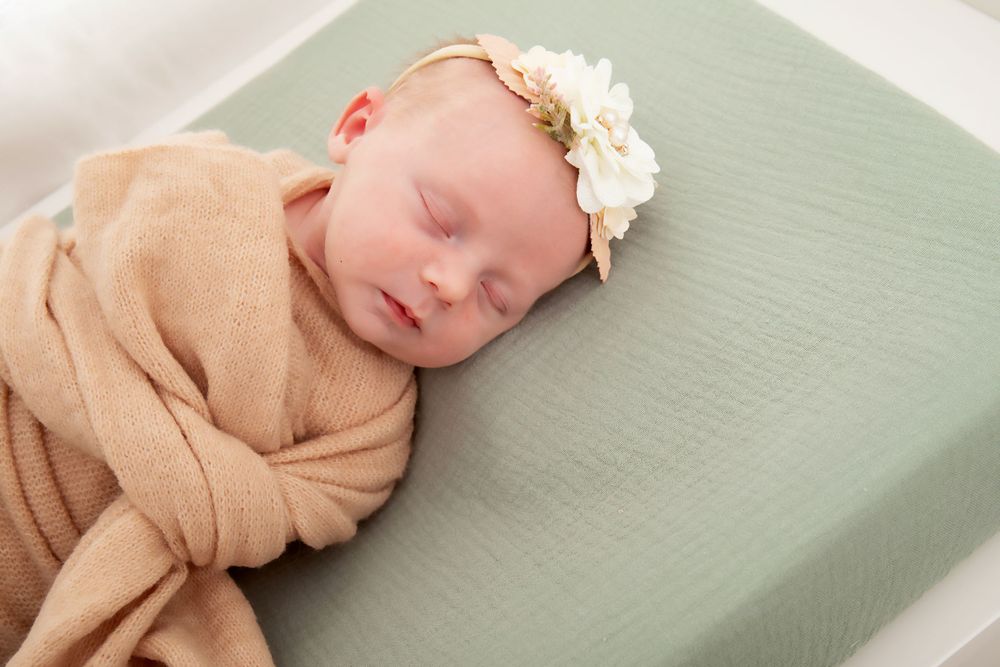 Sleeping baby in beige wrap and floral headband on mint green cushion.