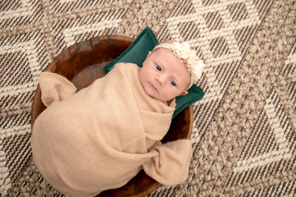 Baby in beige swaddle with floral crown lying on patterned rug with green cushion.