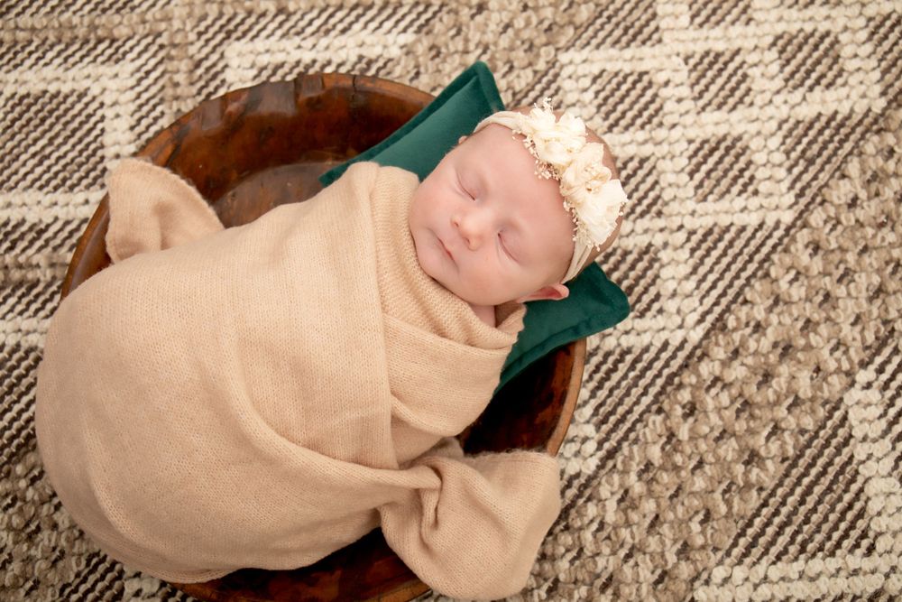 Newborn wrapped in beige blanket resting on brown basket with geometric rug underneath.