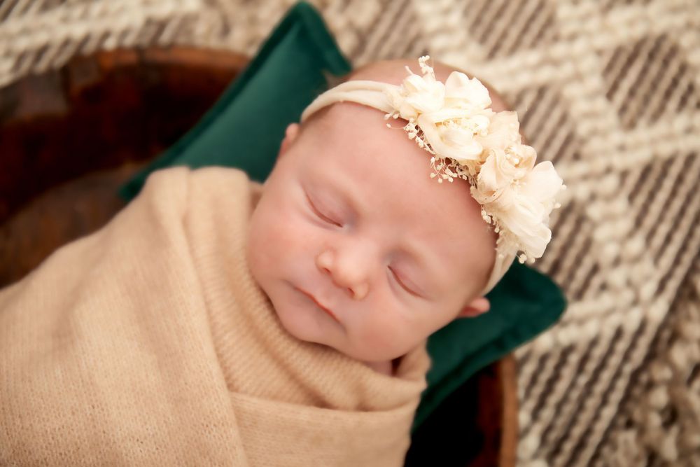 Sleeping newborn wearing delicate floral headband and beige wrap on emerald cushion.