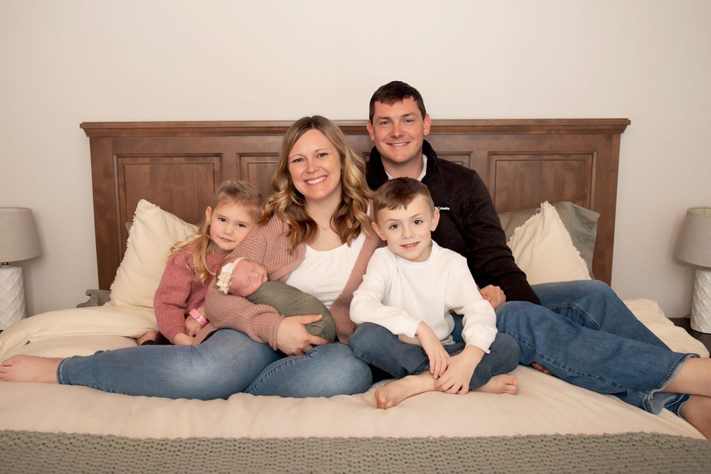 Family lounges together on bed with wooden headboard and cream pillows in casual portrait setting.