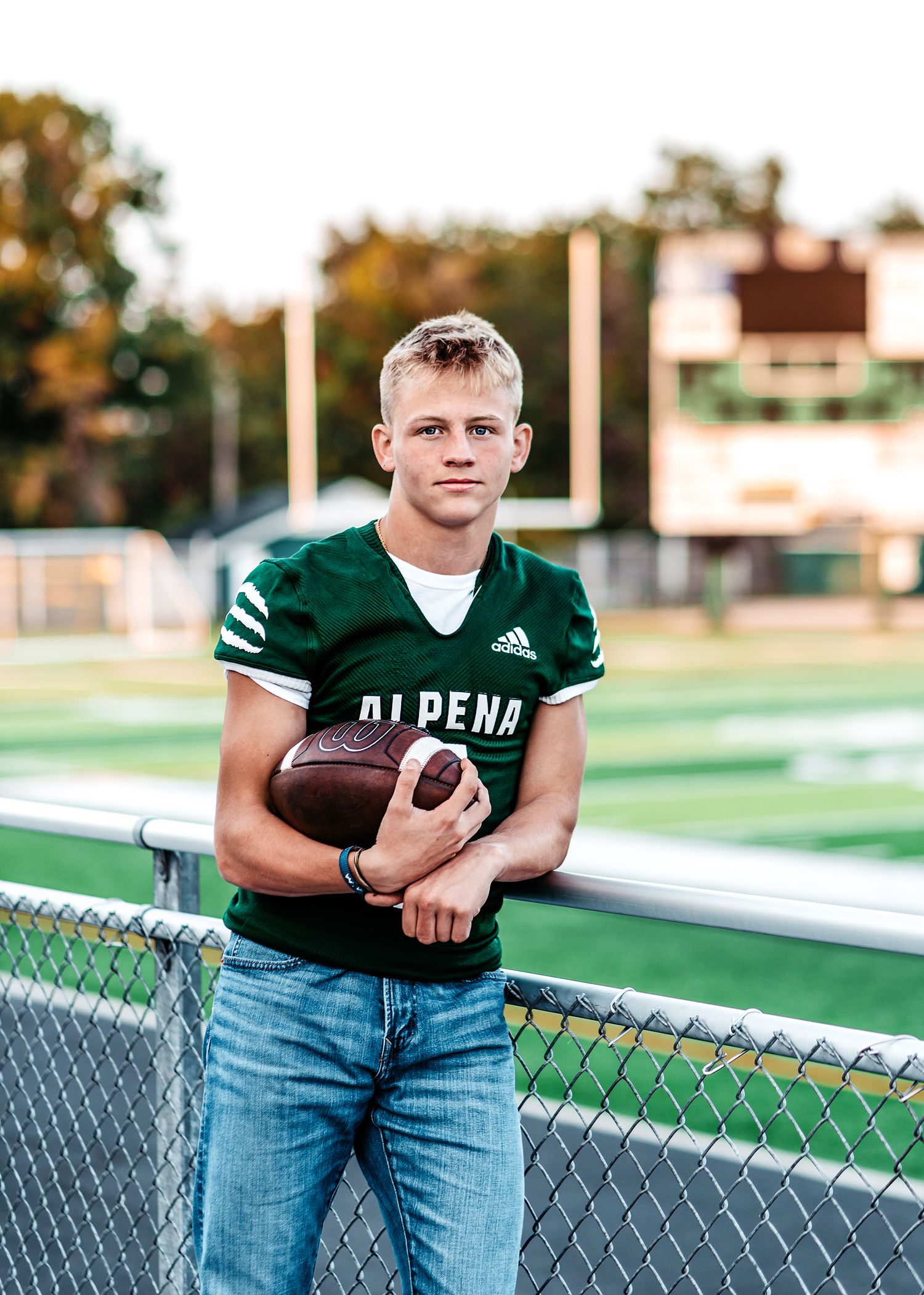 Football player wearing green Alpena jersey and jeans poses with football by field fence at sunset.
