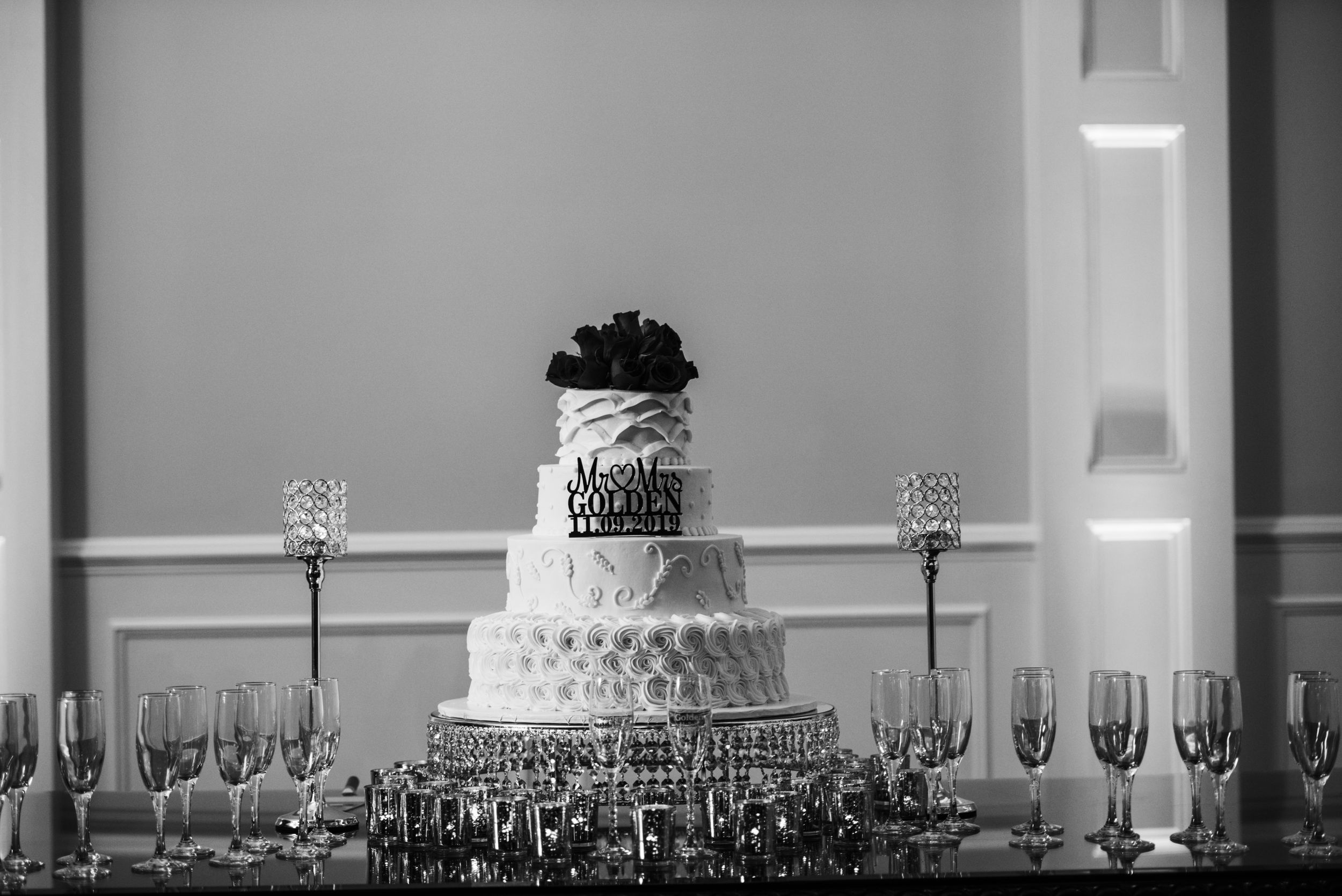 An elegant white wedding cake with detailed patterns sits on a table surrounded by crystal glasses.