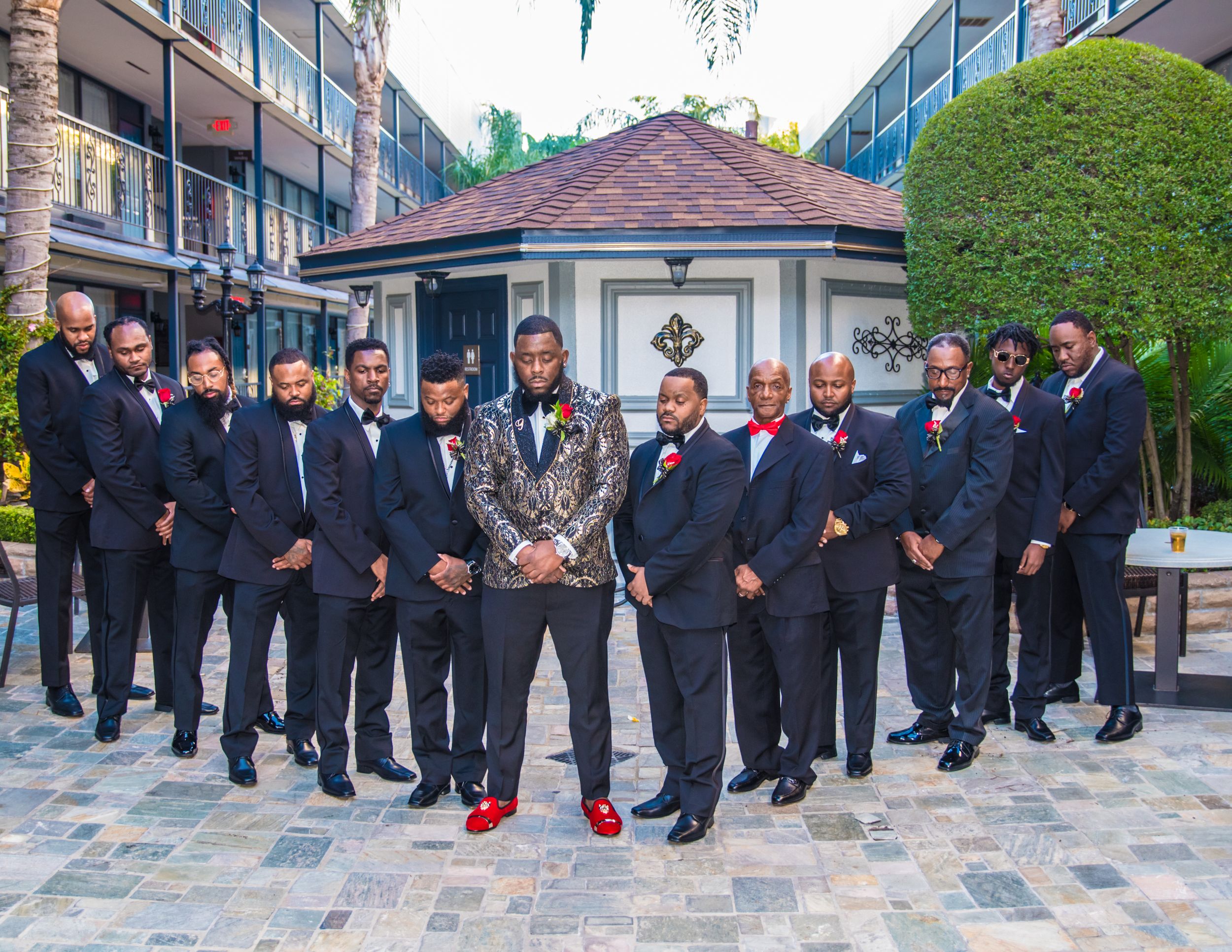 A wedding party of groomsmen in dark suits poses outdoors at an upscale venue with a gazebo.