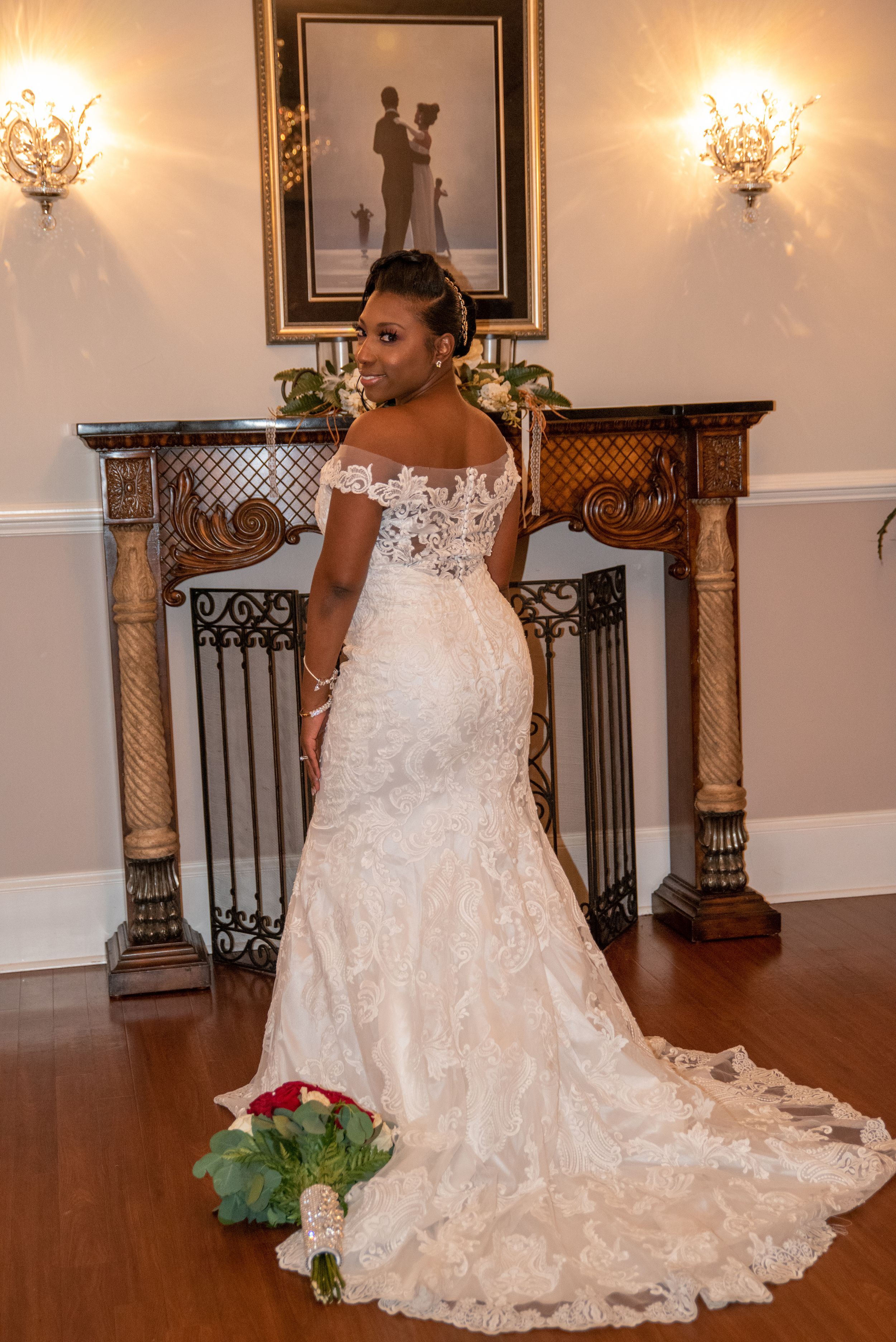 A stunning off-shoulder lace wedding gown with a long train is displayed in front of an elegant fireplace with crystal sconces.