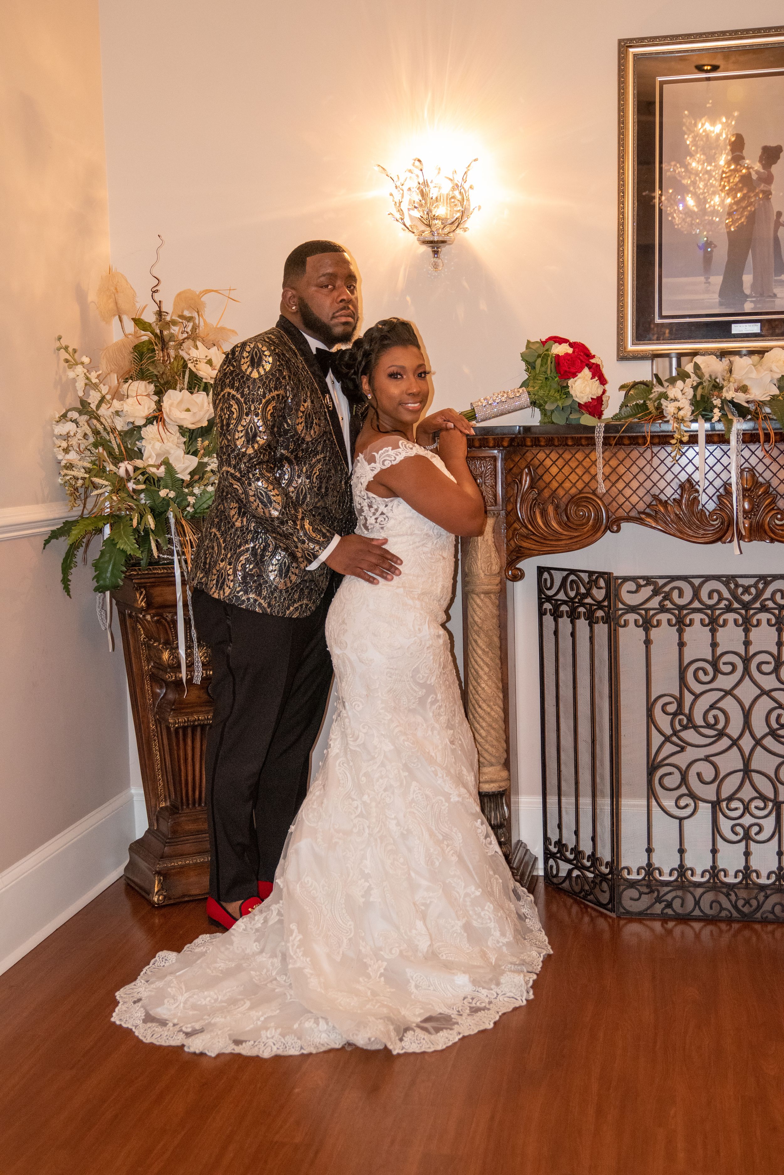 A couple poses in formal wedding attire against an elegant fireplace with floral arrangements.