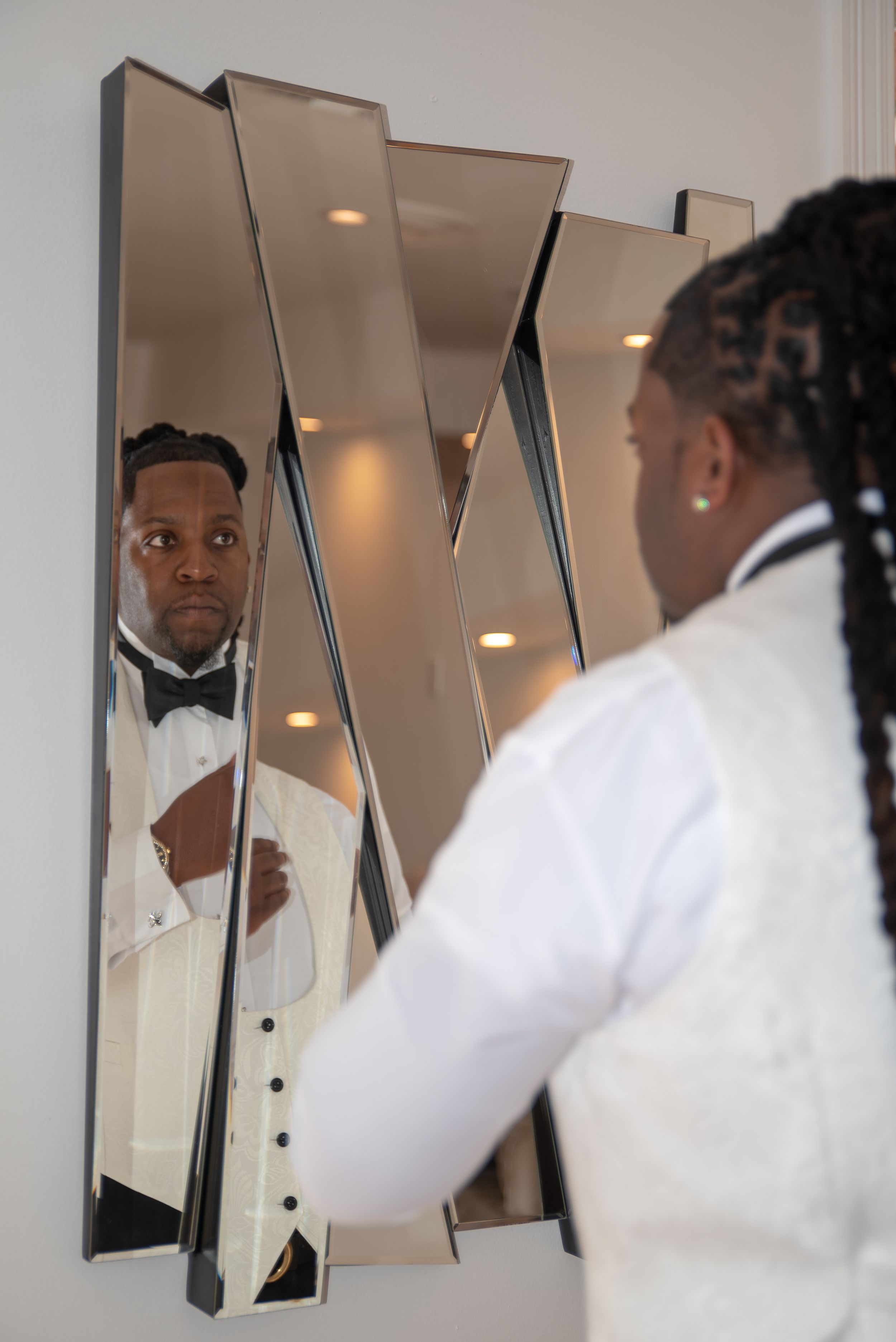 A reflection in geometric mirrors shows formal attire preparation with a white tuxedo jacket and bowtie.