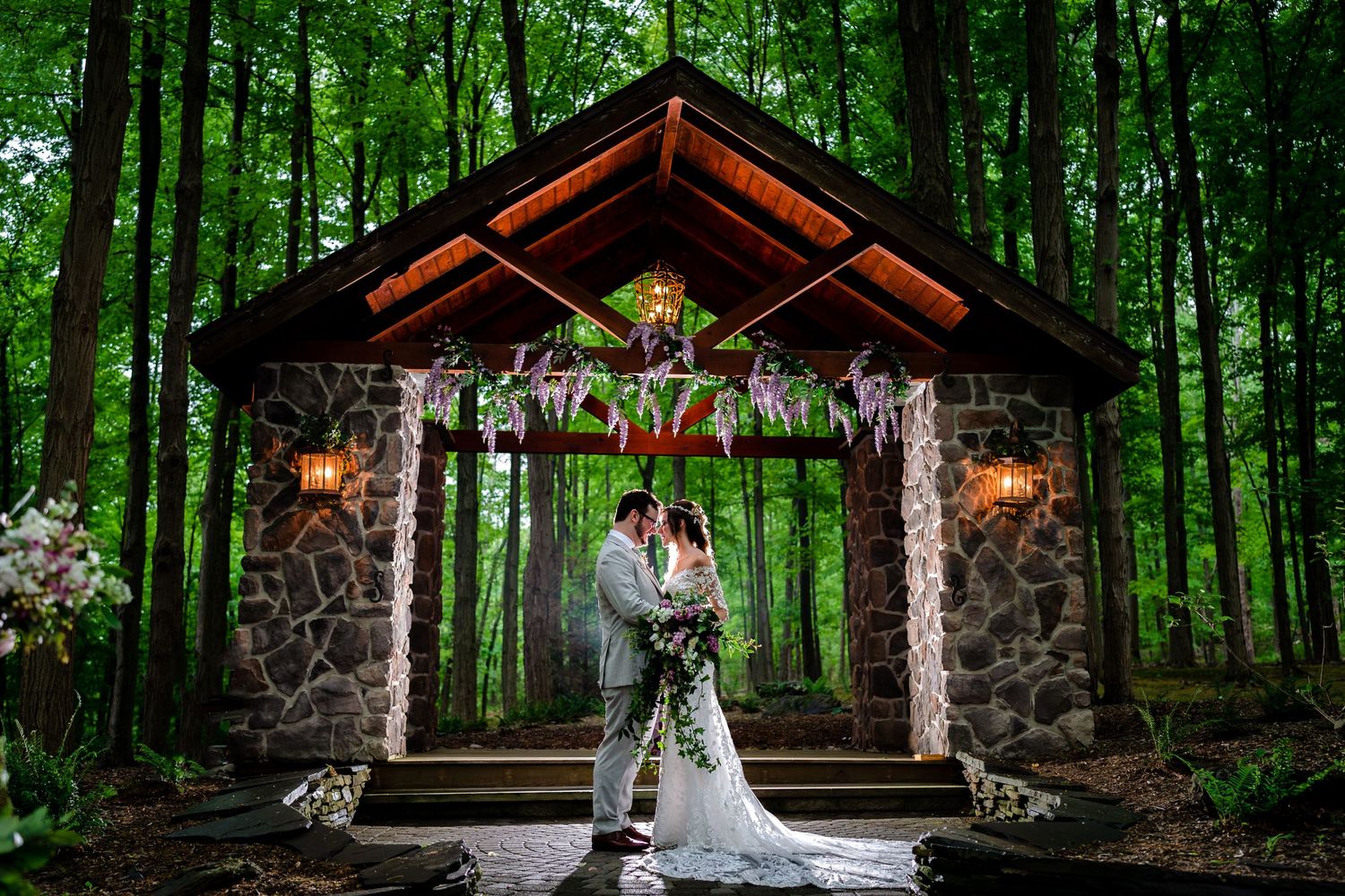 A stone gazebo decorated with flowers and lights creates a magical forest wedding setting.