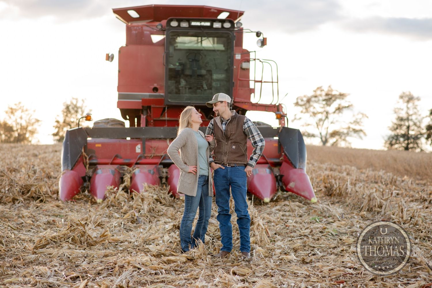 Couple standing together in front of a red combine harvester in a farm field.