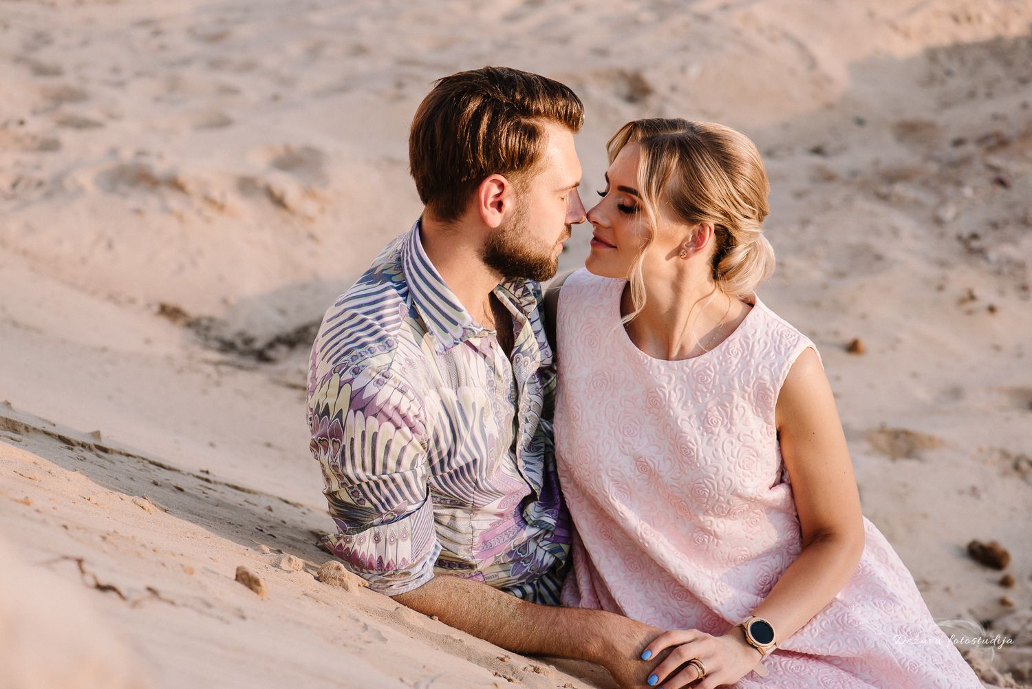 Love story fotosesijos metu sužadėtiniai meiliai žiūri vienas į kitą smėlio kopose