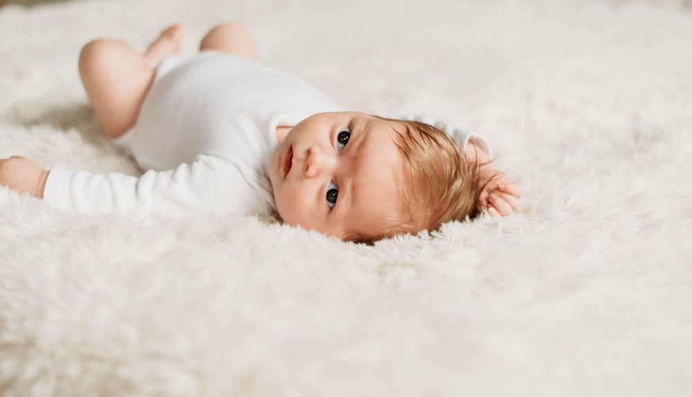 An adorable wide-eyed newborn baby in white onesie lies on a fluffy cream textured rug.