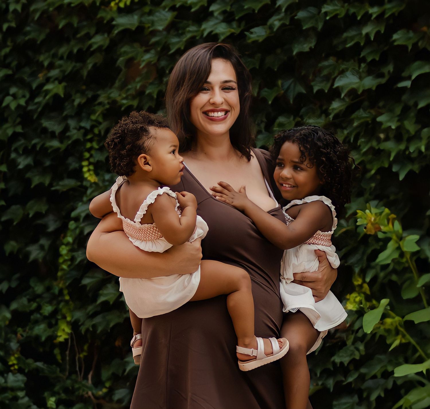 A mother holds two small children in matching outfits against an ivy-covered wall.