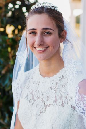 A bride in lace dress and tiara veil smiles in natural lighting.