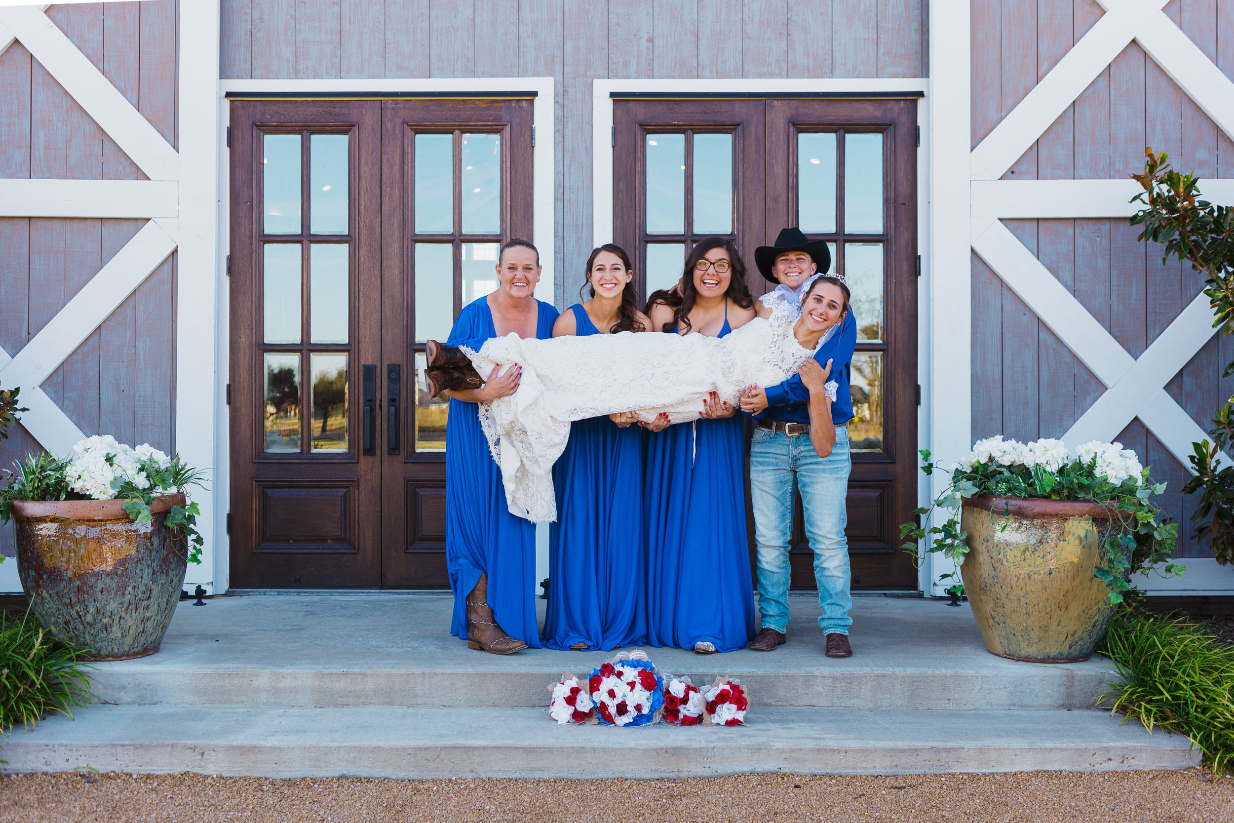 Wedding party members hold a banner while standing on the steps of a rustic barn with blue doors.