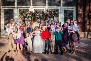 Large group wedding photo with children and adults in front of a floral-decorated building.