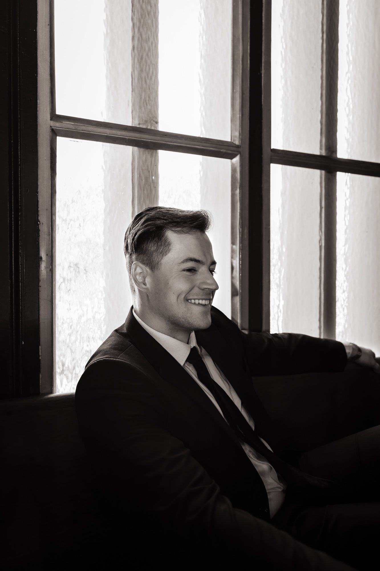 A man sits with his back to a window. He grins and the scene seems very vintage in his suit.