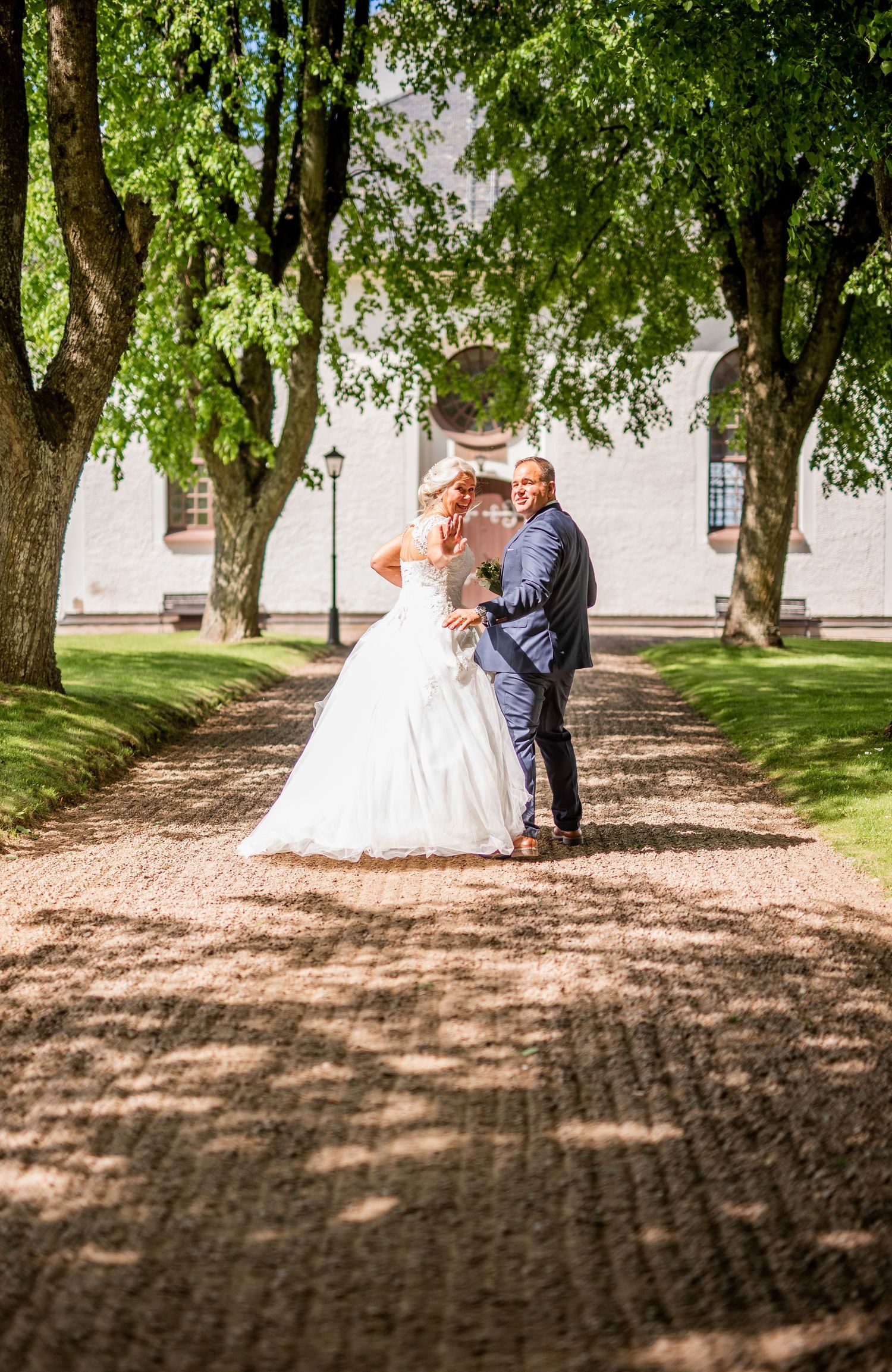 Wedding couple walking on a tree-lined gravel path toward a white building on a sunny day.