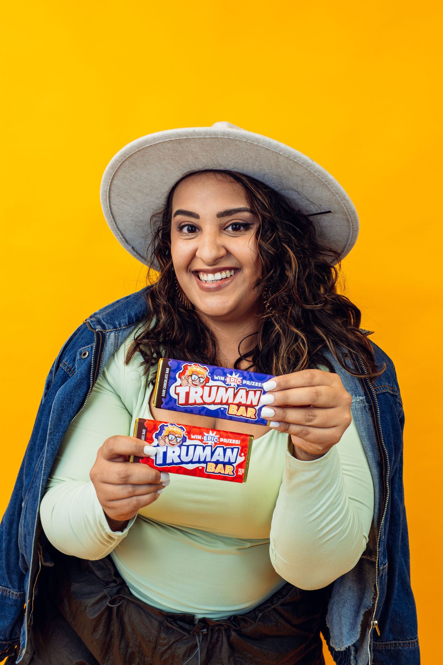 Person in denim jacket and hat displaying Truman candy bars against orange wall.