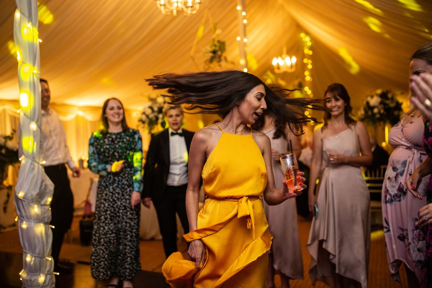 Wedding reception scene in a yellow-lit tent with dancers celebrating on the dance floor.