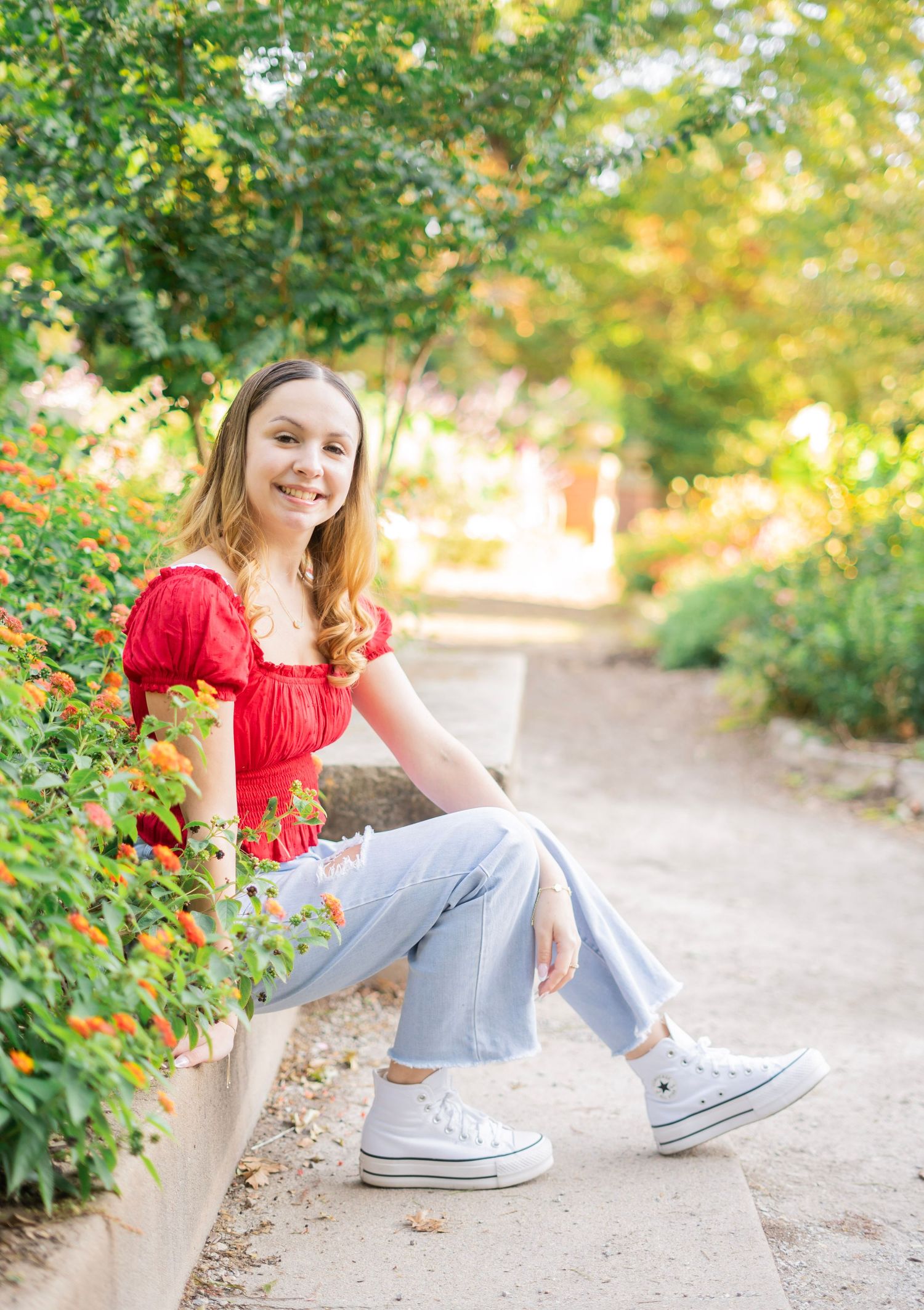 Young woman in red top and jeans sitting by orange flowers on a garden path.