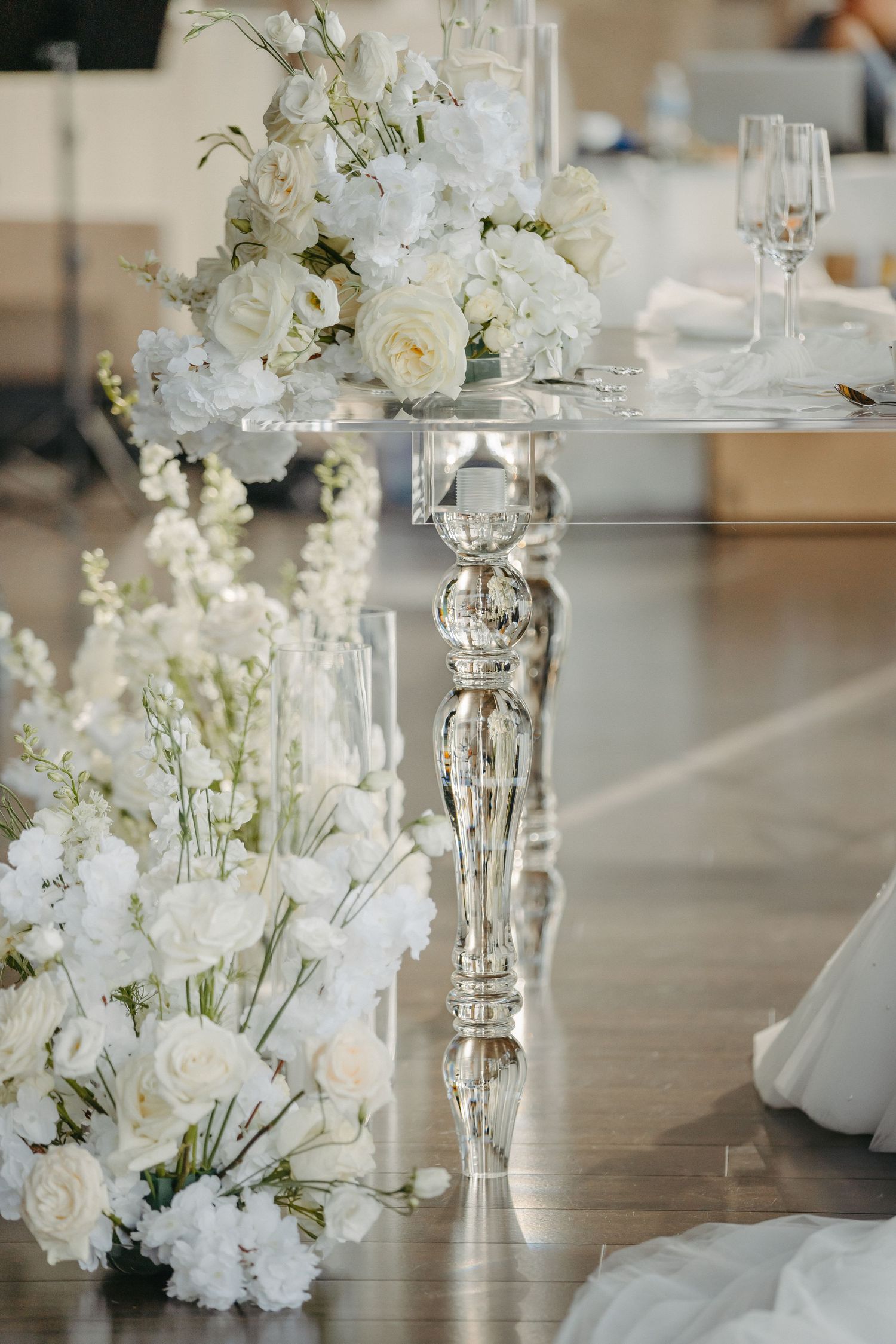 Elegant white floral centerpiece with roses on a crystal pedestal table with champagne glasses.