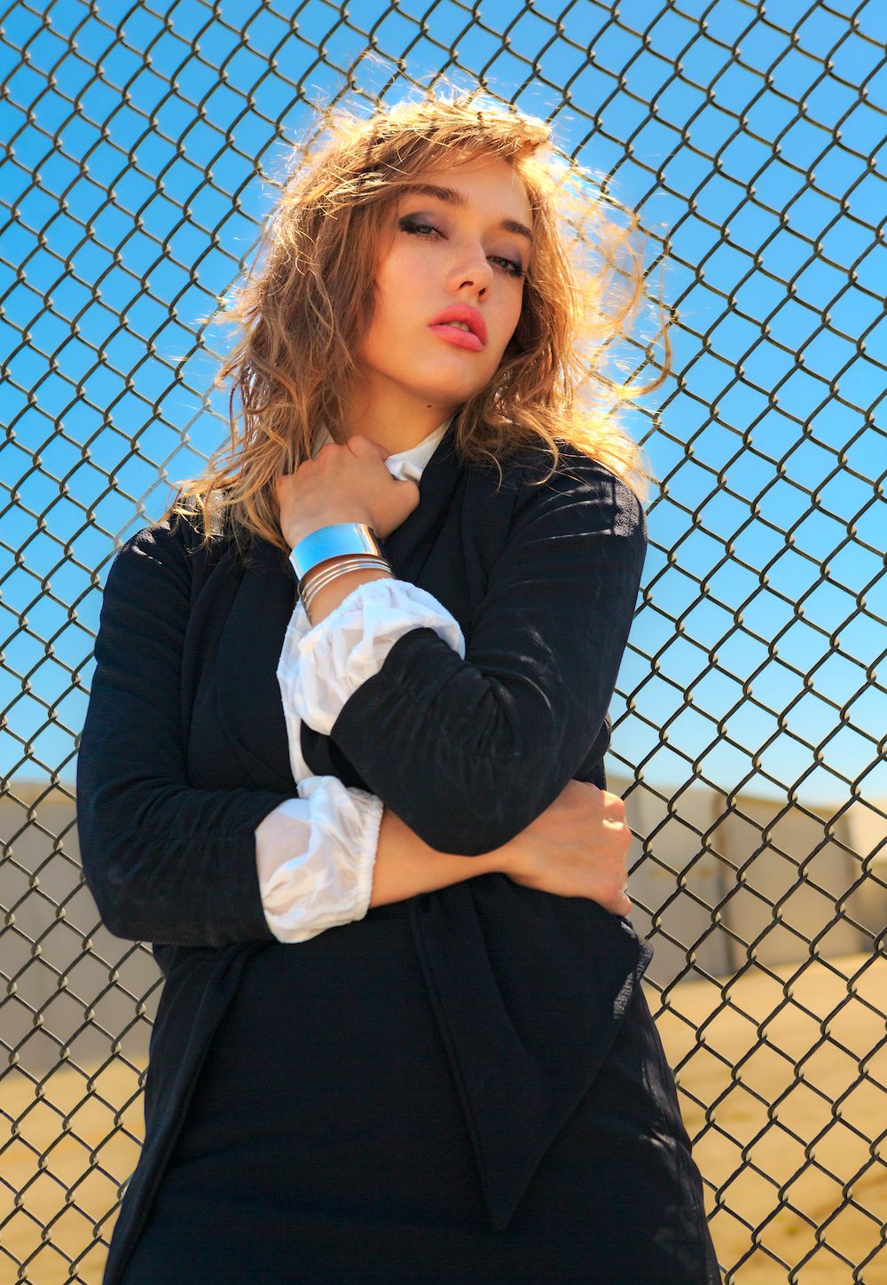 Colorful, outdoor high-fashion image of blonde model in a black jacket, white shirt and silver jewelry, in front of a chainlink fence.