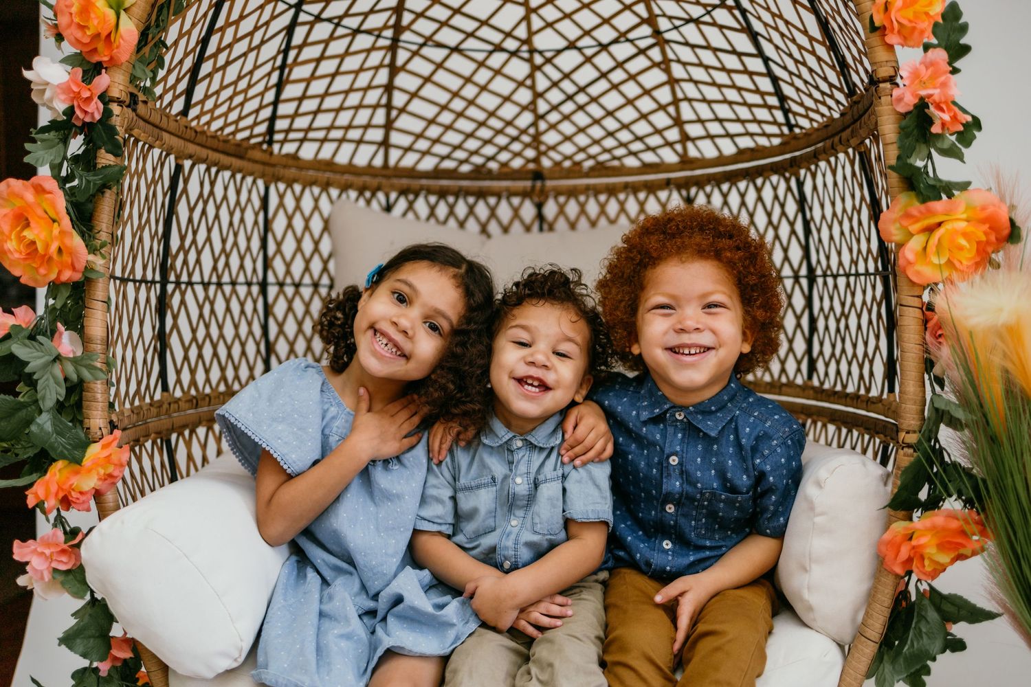 Three happy children sitting together in a wicker egg chair decorated with colorful flowers.
