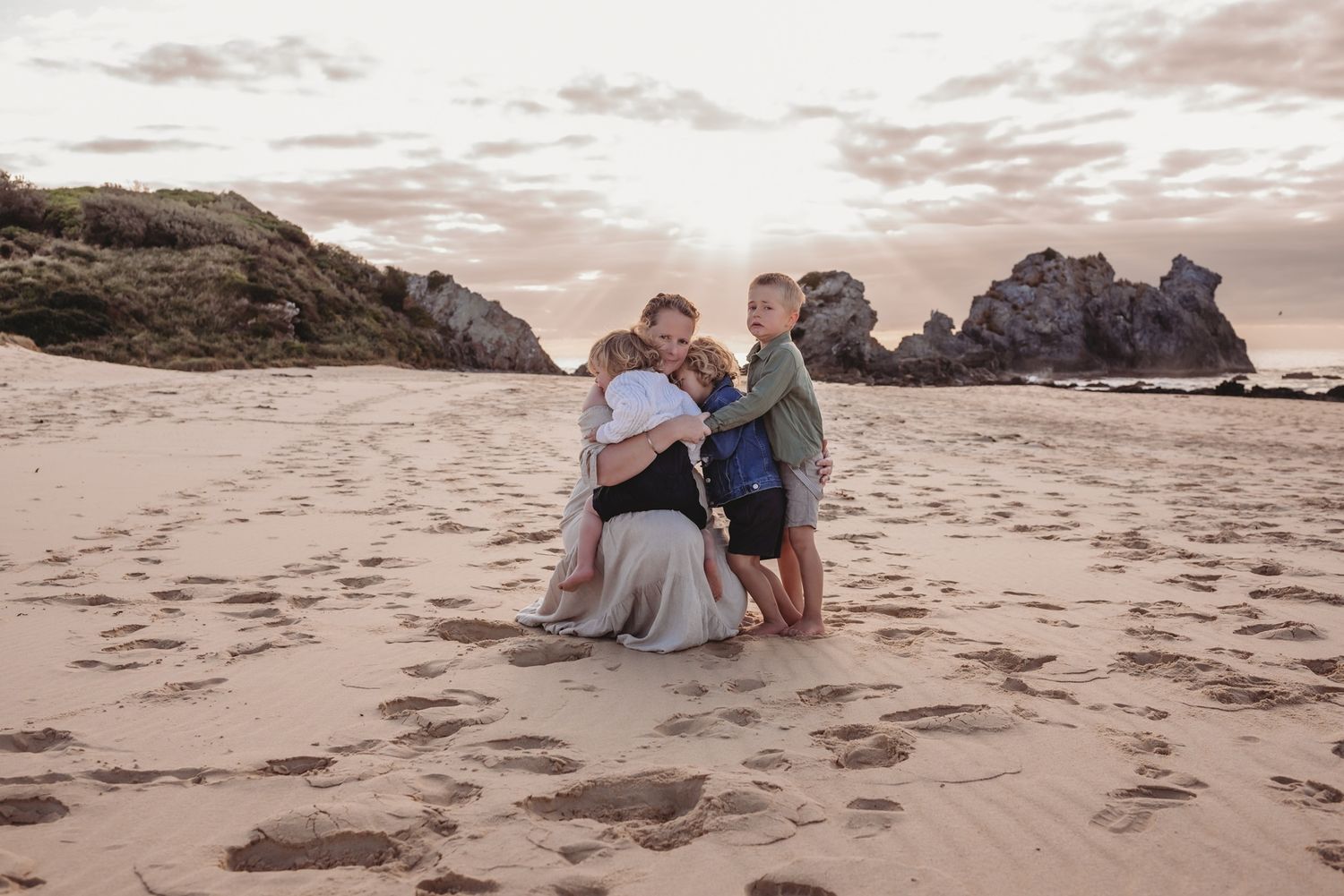 Family embraces on sandy beach at sunset with rocky cliffs in background.
