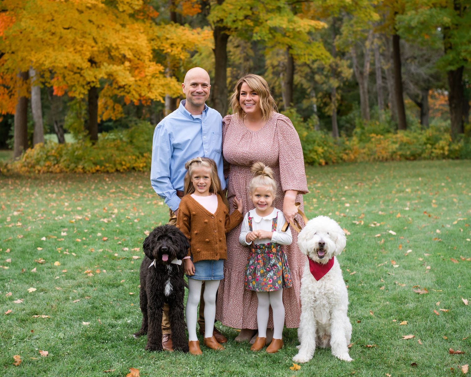 A family with two children and three dogs pose together on grass with autumn foliage in the background.