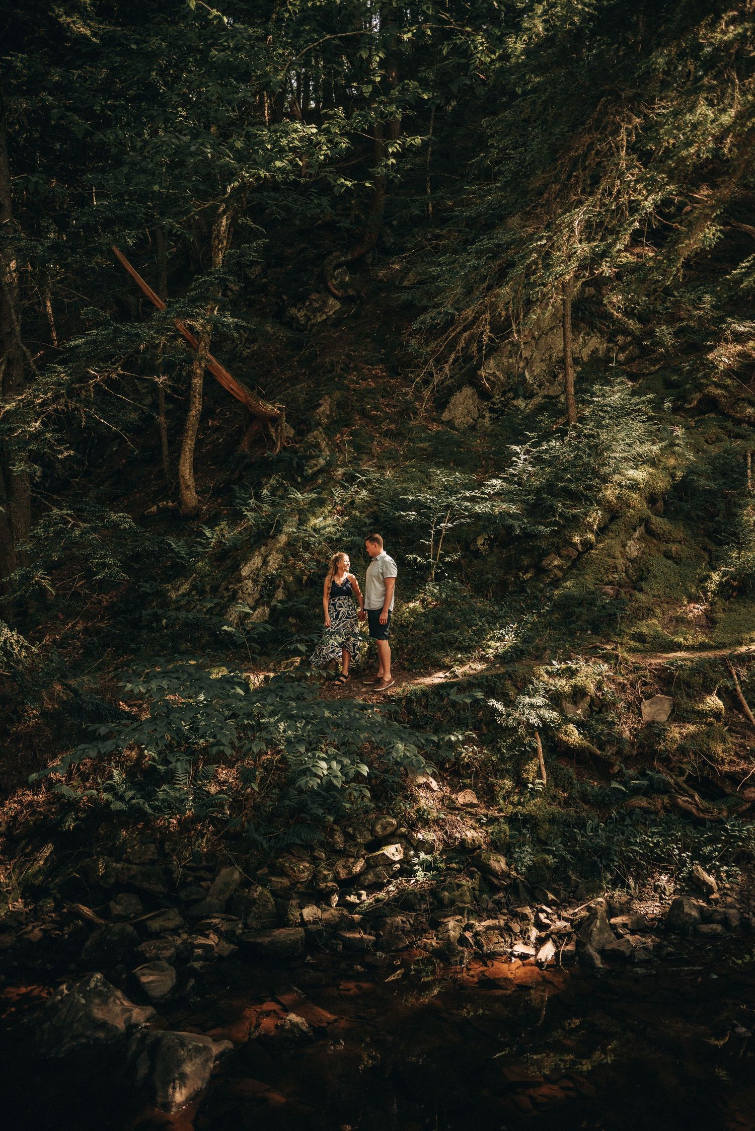 A couple hiking through a lush forest trail surrounded by towering trees and rocky terrain.