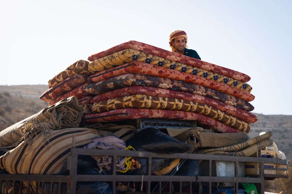 Stack of colorful traditional carpets and textiles loaded on top of a vehicle in a desert setting.