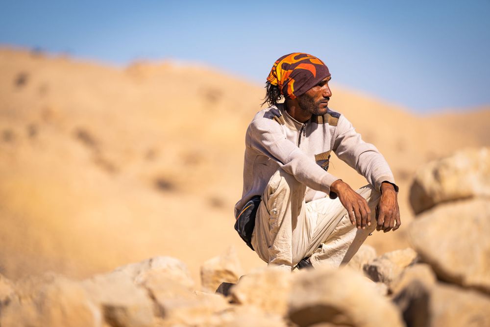Desert dweller in traditional clothing resting on rocky terrain under bright blue sky.