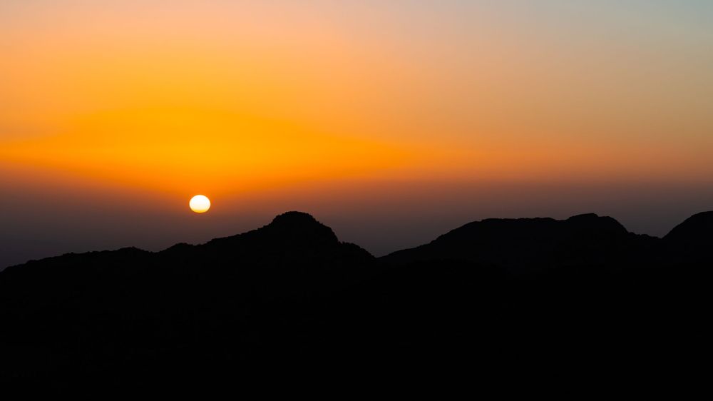 Setting sun creates silhouette of mountain peaks against vibrant orange desert sky.
