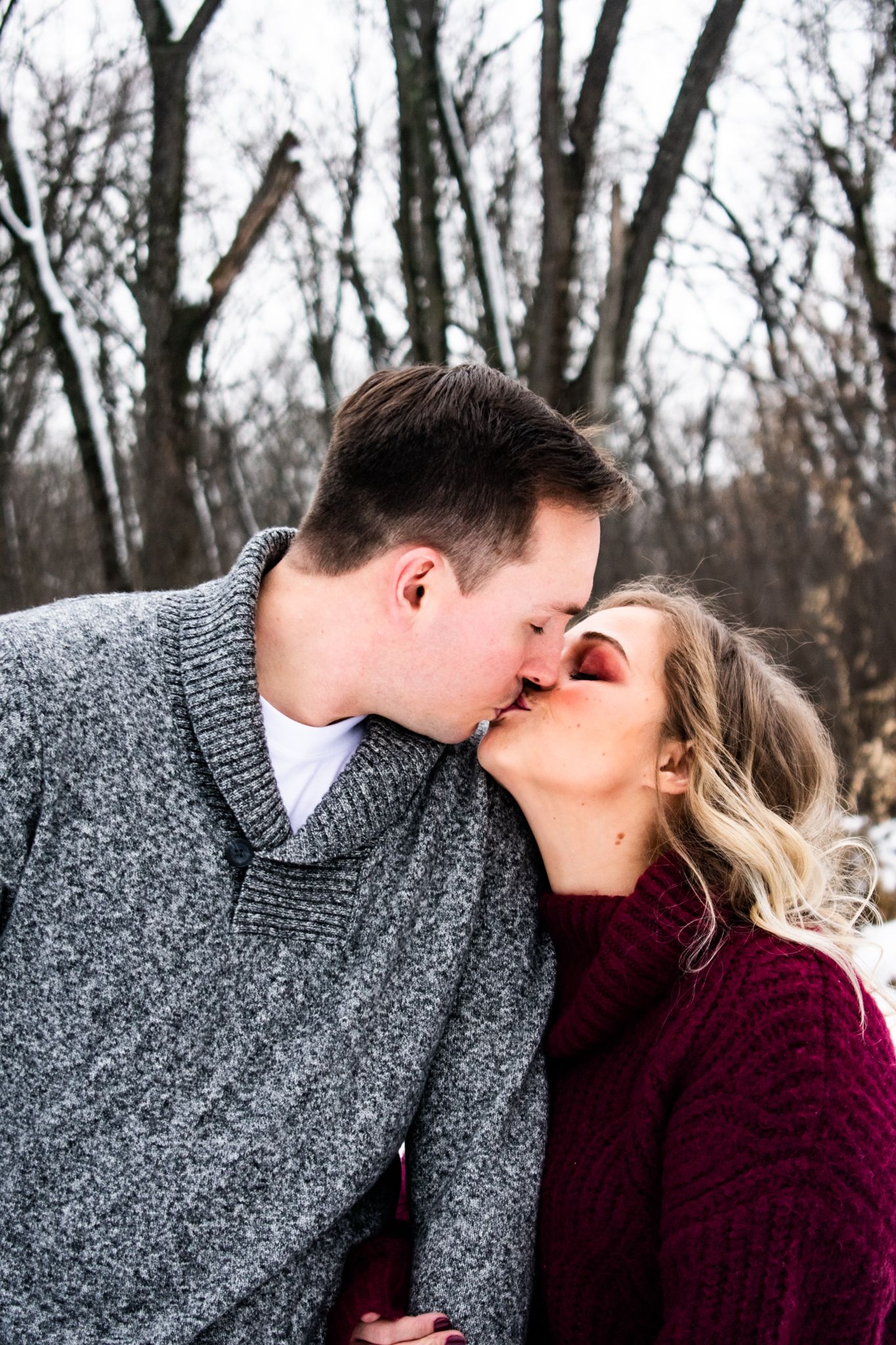 A romantic winter kiss captured between two people wearing cozy sweaters among bare trees.