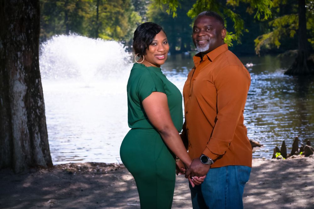 A couple in orange and green outfits pose together by a lake in a natural setting.