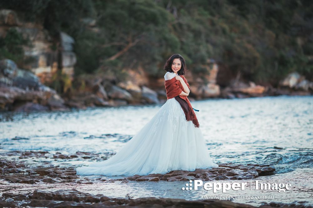 Bride in white tulle gown and rust wrap posing on rocky waterfront surrounded by trees.