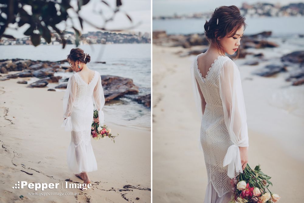 Elegant bride in white lace gown holding bouquet on sandy beach, back to camera.