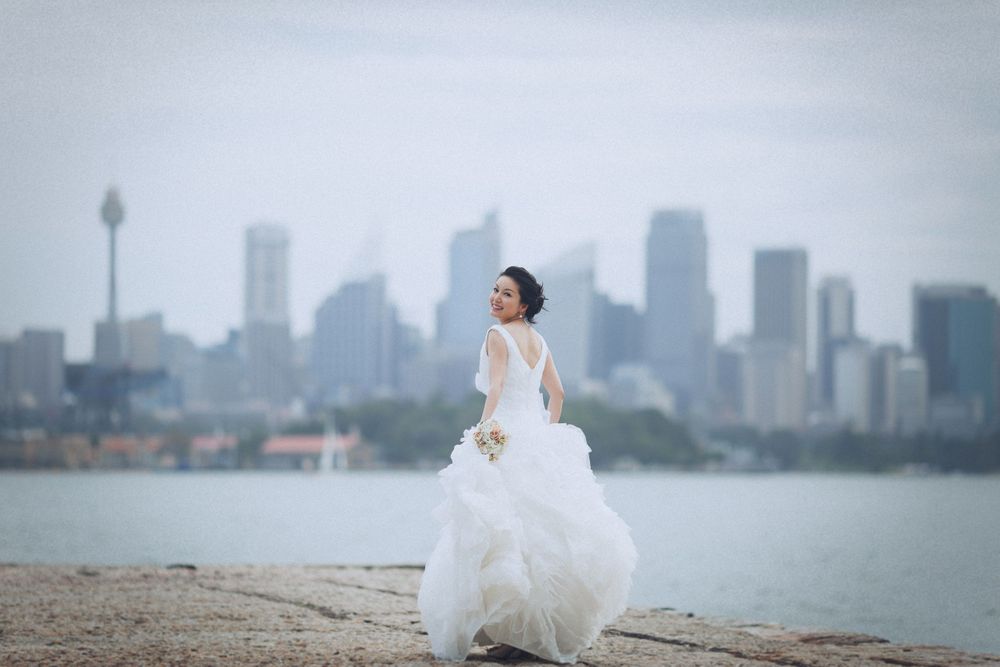 Bride in flowing white gown twirling on waterfront with Sydney city skyline behind her.