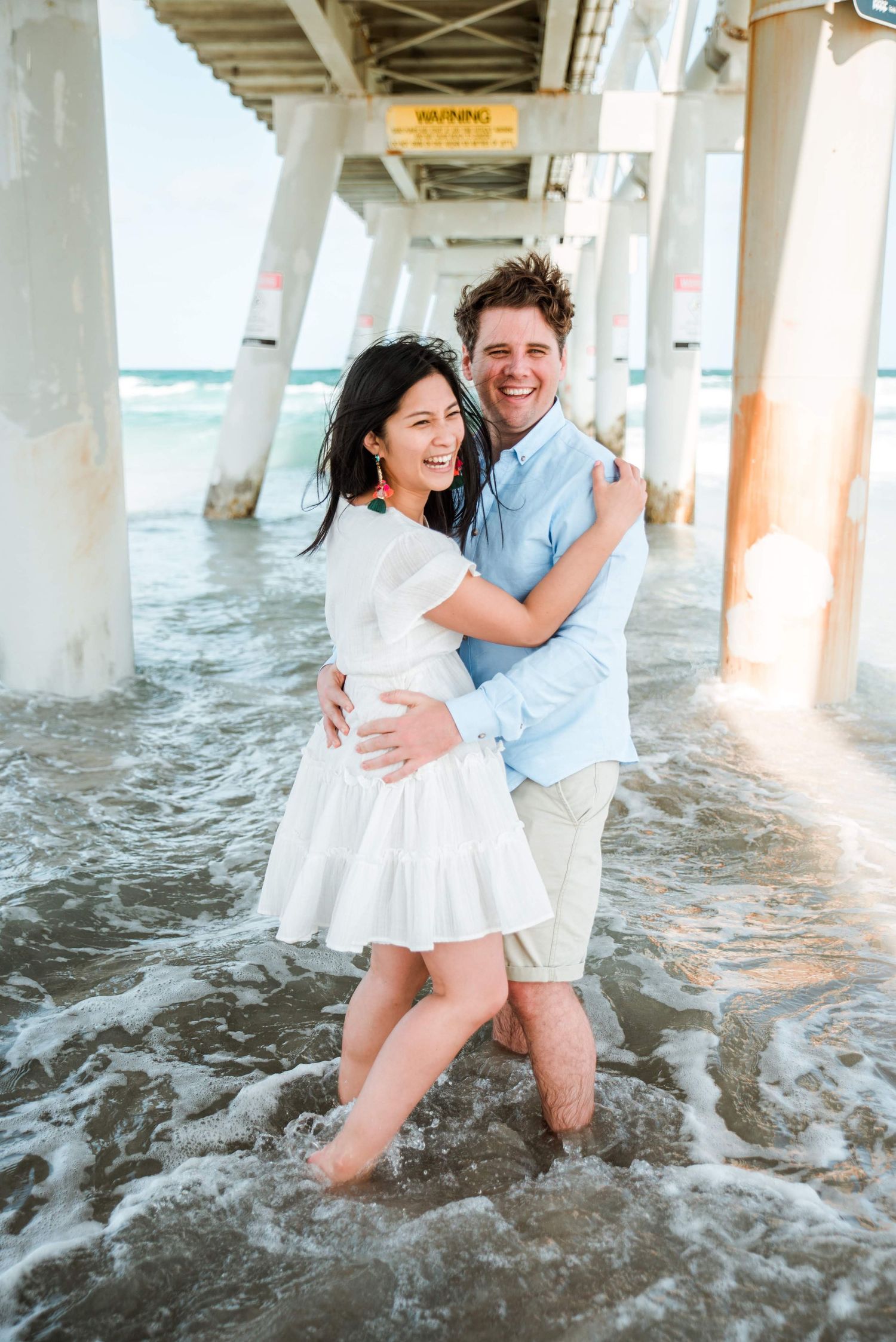 A couple stands together in the waves beneath a wooden pier, embracing and smiling.