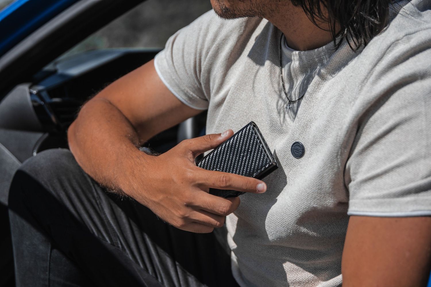 Person in white t-shirt holding carbon fiber wallet while sitting in car.